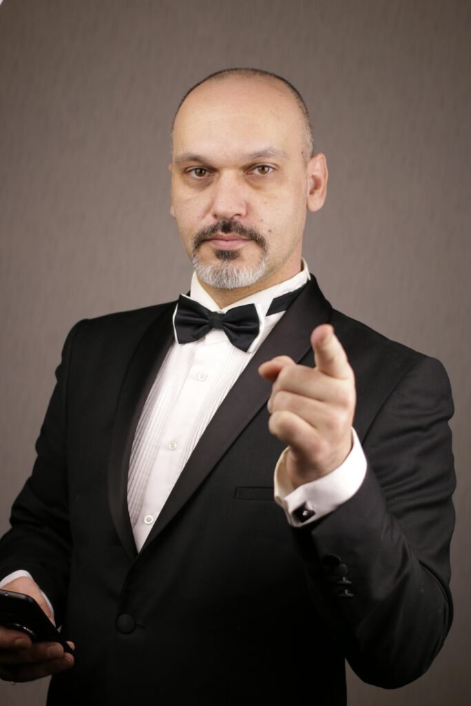 Portrait of a confident bald man in a tuxedo pointing at the camera in a studio setting.